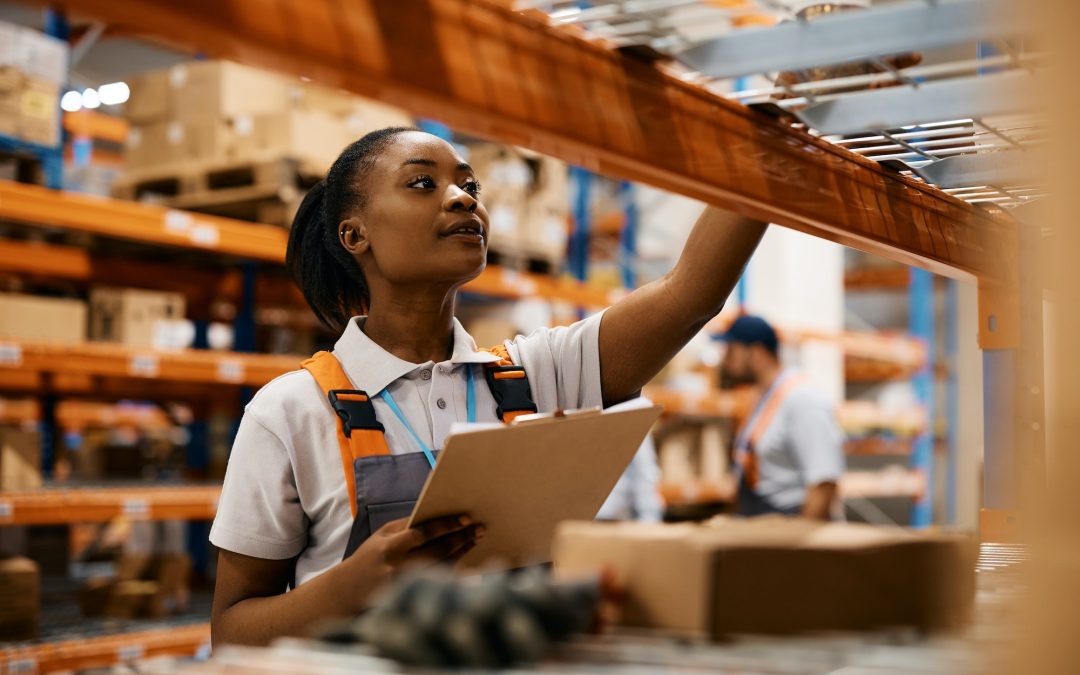 Black female worker checking inventory on shelves of distribution warehouse.