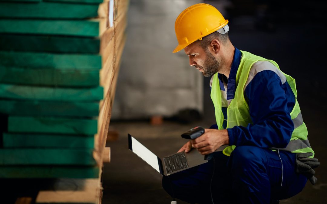 Warehouse worker using laptop and bar code scanner at lumber storage compartment.